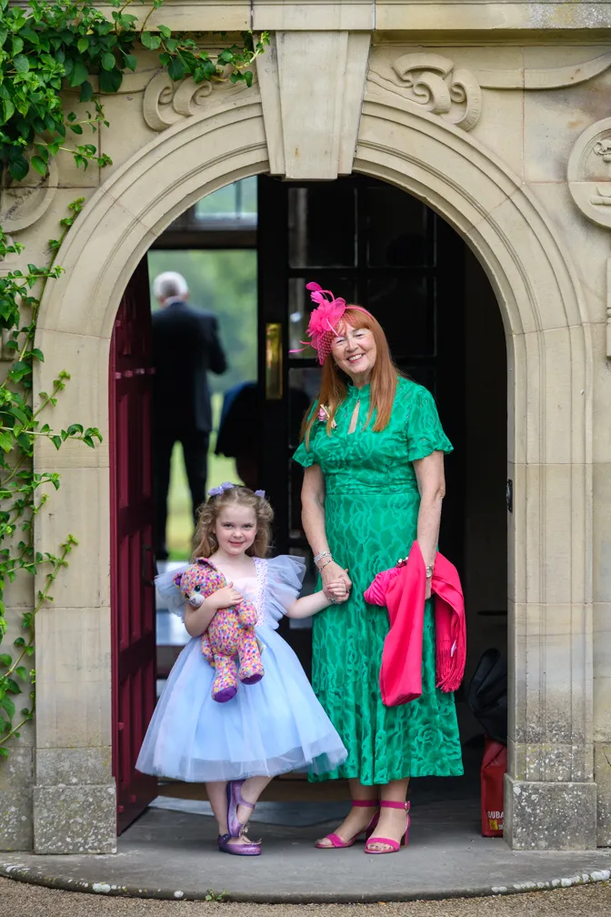 A smiling woman in a green dress and pink fascinator holding hands with a young girl in a blue dress holding a colorful stuffed bear, standing under an ornate stone archway.