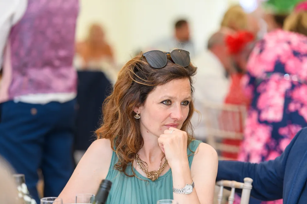 Thoughtful woman with sunglasses on her head, wearing a teal dress and gold necklace, sitting at a table at a social event.