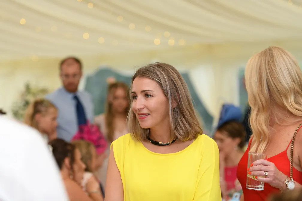 Woman in a yellow dress smiling at an indoor event with other guests blurred in the background.