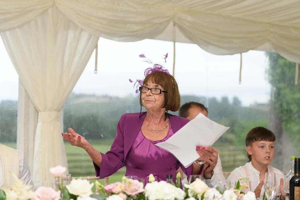 An elderly woman in a purple dress and fascinator giving a speech while holding papers at a decorated table.