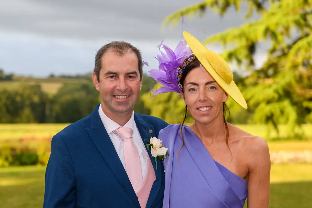 Man in a blue suit with a light pink tie and boutonniere standing next to a woman wearing a one-shoulder purple dress and a large yellow hat with purple feathers.