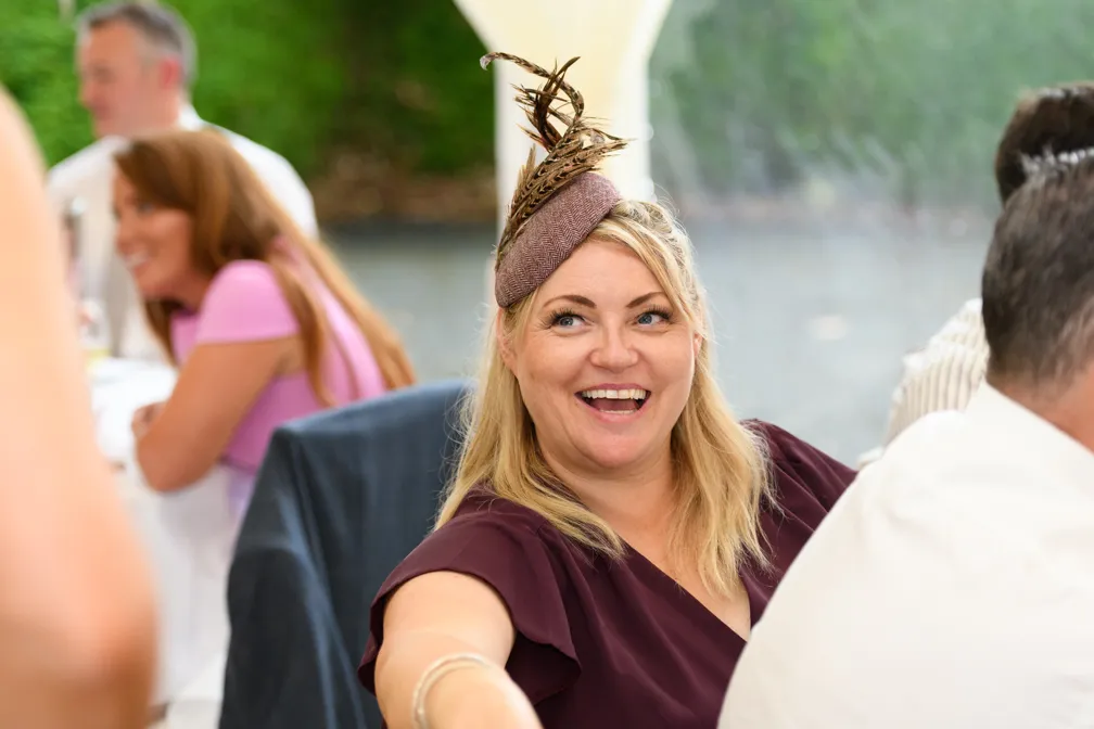 Smiling woman with blonde hair wearing a feathered fascinator and burgundy dress at a social event.