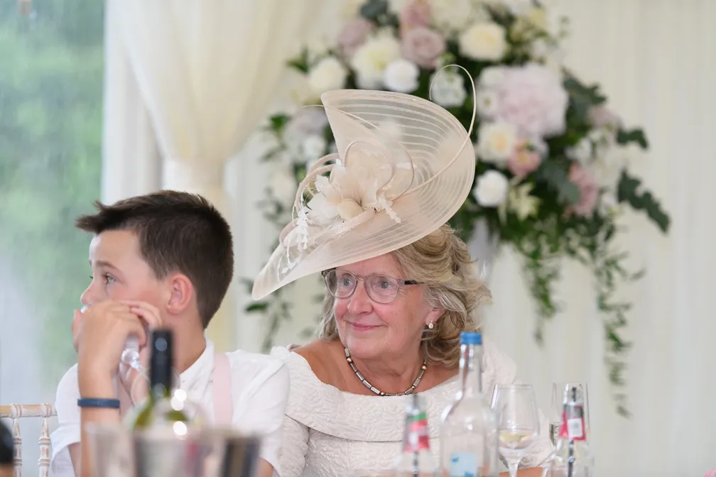 Older woman wearing a large beige floral hat and glasses seated at a table next to a young boy in white shirt, with a floral arrangement in the background.