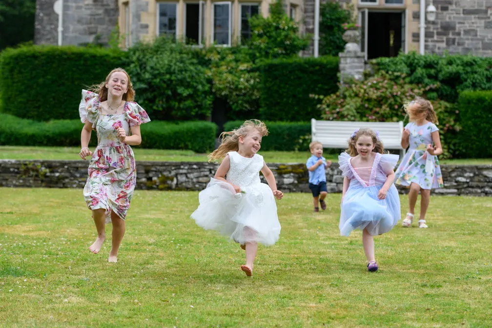 Five children in colorful dresses and a shirt running and playing on a green lawn in front of a stone house with greenery.