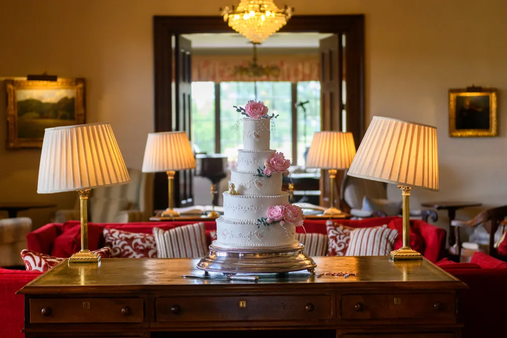 Four-tier white wedding cake decorated with pink flowers on a wooden table with brass lamps and red upholstered furniture in the background.