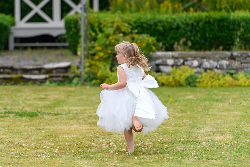 Young girl in a white dress with a large bow running barefoot on grass in a garden.