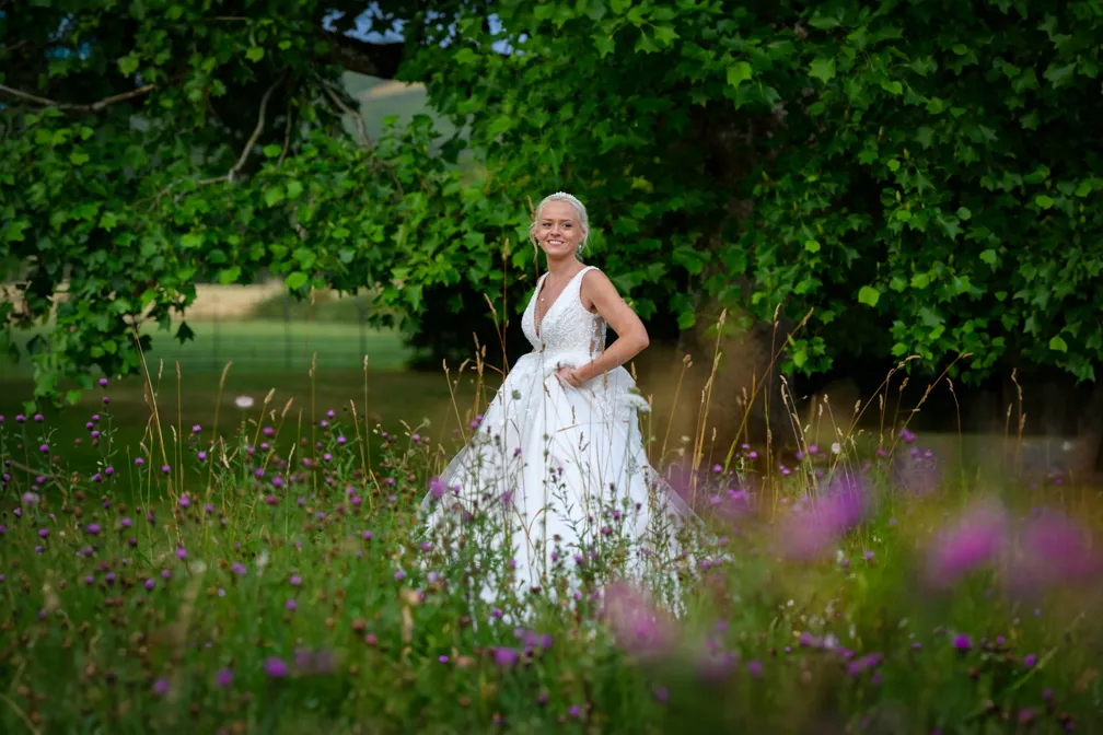 Smiling bride in a white sleeveless wedding gown standing in a wildflower meadow with large green trees in the background.