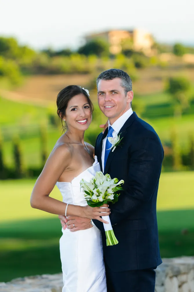 Bride in white strapless gown holding a bouquet and groom in dark suit with blue tie embracing outdoors.