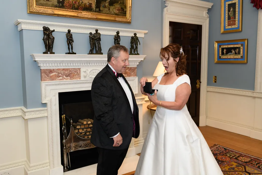 Bride in white gown showing a small black box to a man in a black tuxedo, standing in an elegant room with a fireplace and statues.
