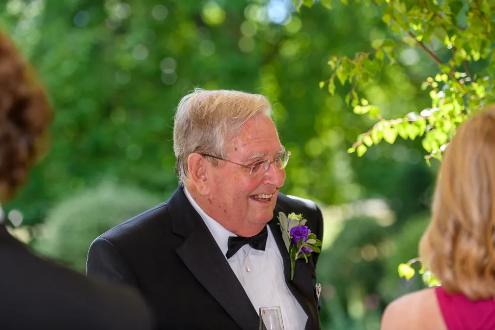 Elderly man in a tuxedo with a purple boutonniere smiling outdoors at a social event.