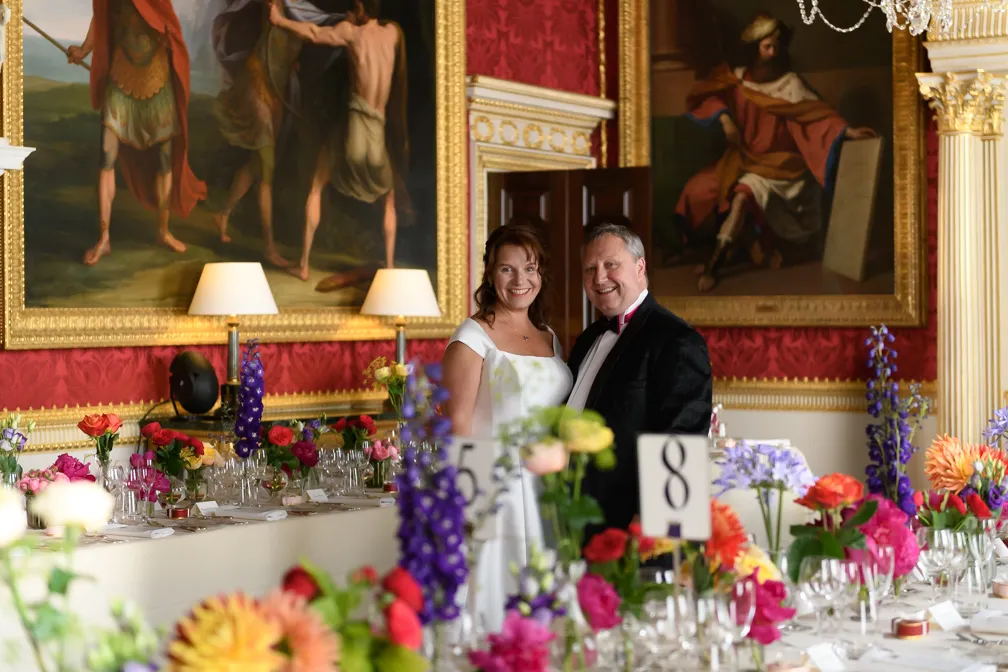 Bride in white dress and groom in black tuxedo smiling in an ornate room with red wallpaper, floral-decorated tables, and large framed paintings.