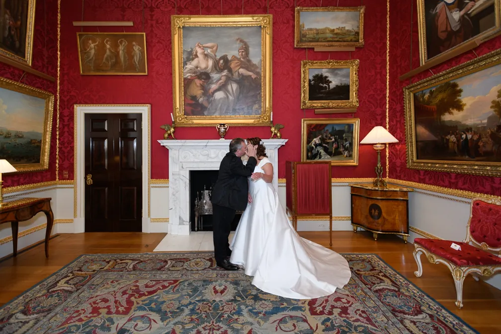 Bride in white wedding gown and groom in black suit sharing a kiss in a richly decorated room with red walls, gold-framed paintings, and a patterned rug.
