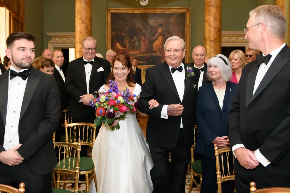 Bride in white dress holding colorful bouquet, walking arm in arm with an older man in a tuxedo, surrounded by formally dressed guests in a decorated room.