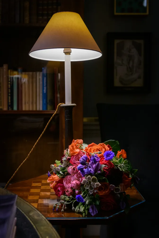 Table lamp with beige shade illuminating a colorful bouquet of mixed flowers on a wooden chessboard table.