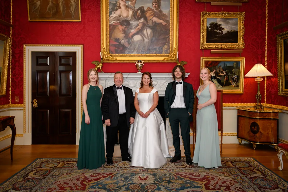 Group of five people dressed in formal attire standing in an ornate room with red wallpaper, framed paintings, and a decorative fireplace.
