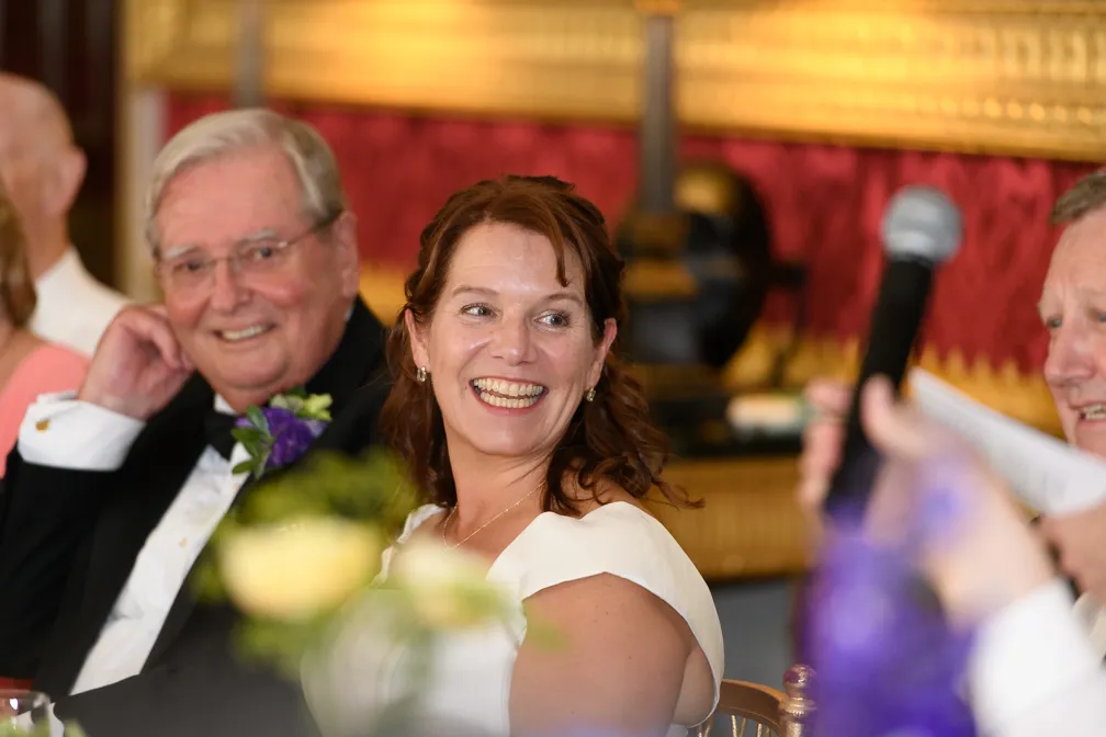 Woman in white dress smiling while seated next to an older man in a tuxedo with a purple boutonniere during an event speech.