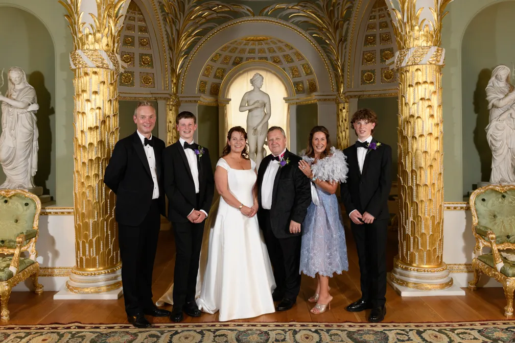 Group of six people dressed in formal attire posing in a grand room with golden ornate columns and classical statues in the background.