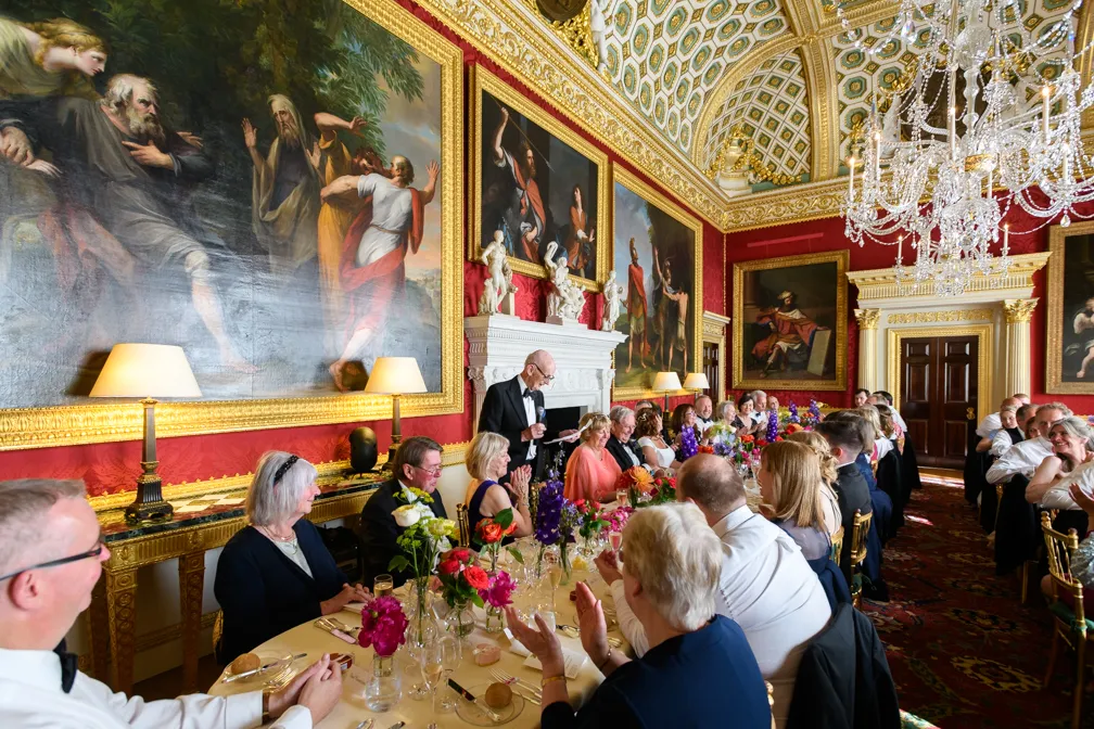 Elegant formal dinner with guests seated along a long table in a luxurious room featuring large classical paintings, ornate gold decor, and a crystal chandelier.