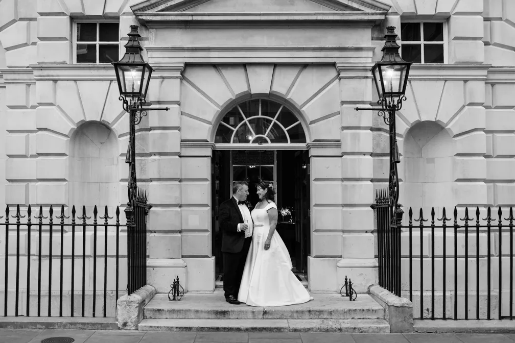Bride and groom standing on steps outside a grand building with classic stone architecture and wrought iron railings.