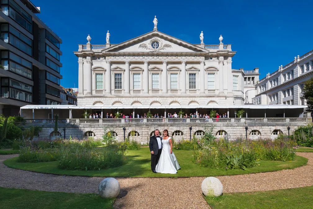 Bride and groom standing on garden path in front of a large historic white building with guests on the balcony.