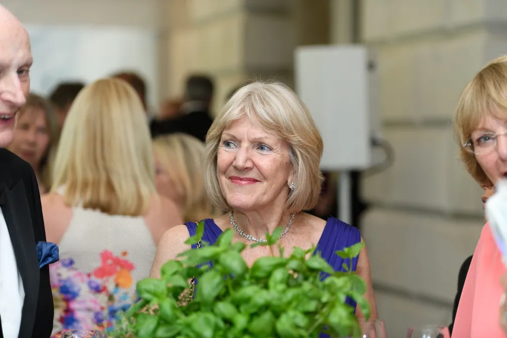 Smiling older woman in a blue dress with silver necklace, holding green leafy plants at a social gathering.
