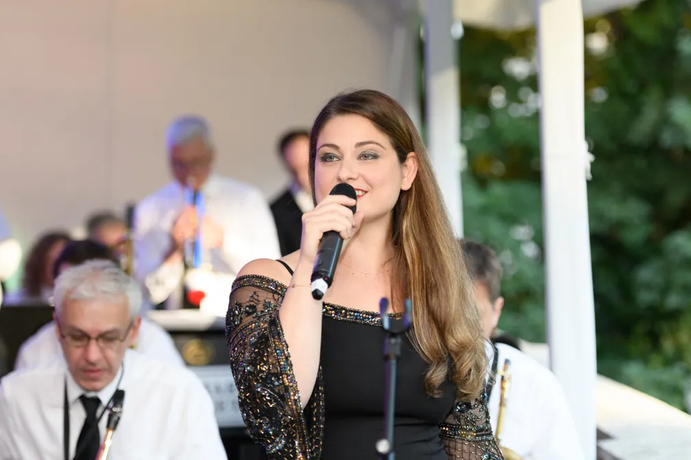 A woman with long brown hair in a black, beaded off-shoulder dress singing into a microphone, with musicians playing instruments in the background.