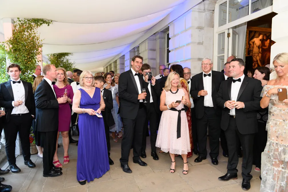 Group of elegantly dressed people in formal attire at a social event, standing under a white canopy outside a building.