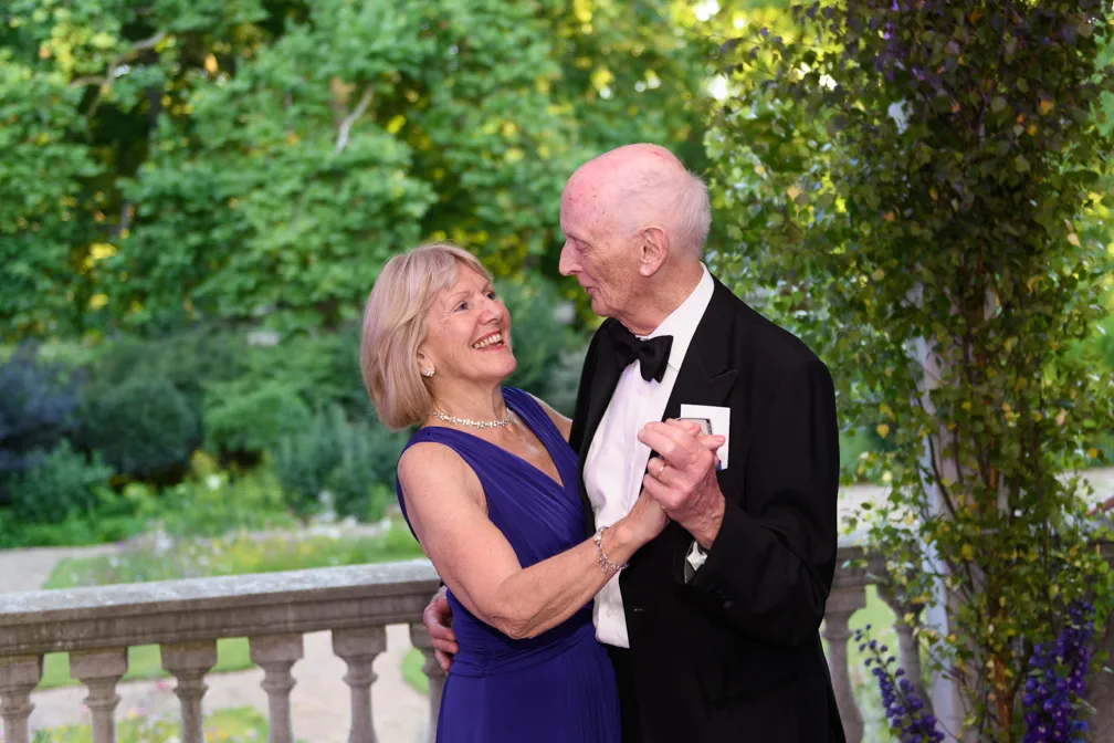 Elderly couple dressed formally dancing outdoors near a stone balustrade with green foliage background.