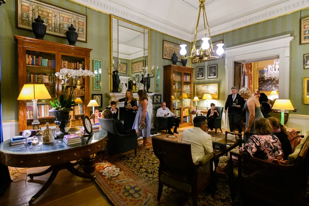 A group of people socializing in an elegantly furnished living room with vintage decor, bookshelves, lamps, and a chandelier.