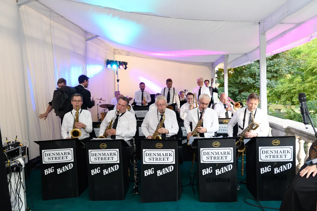 Denmark Street Big Band performing under a white canopy tent with musicians playing saxophones, trombones, and drums.