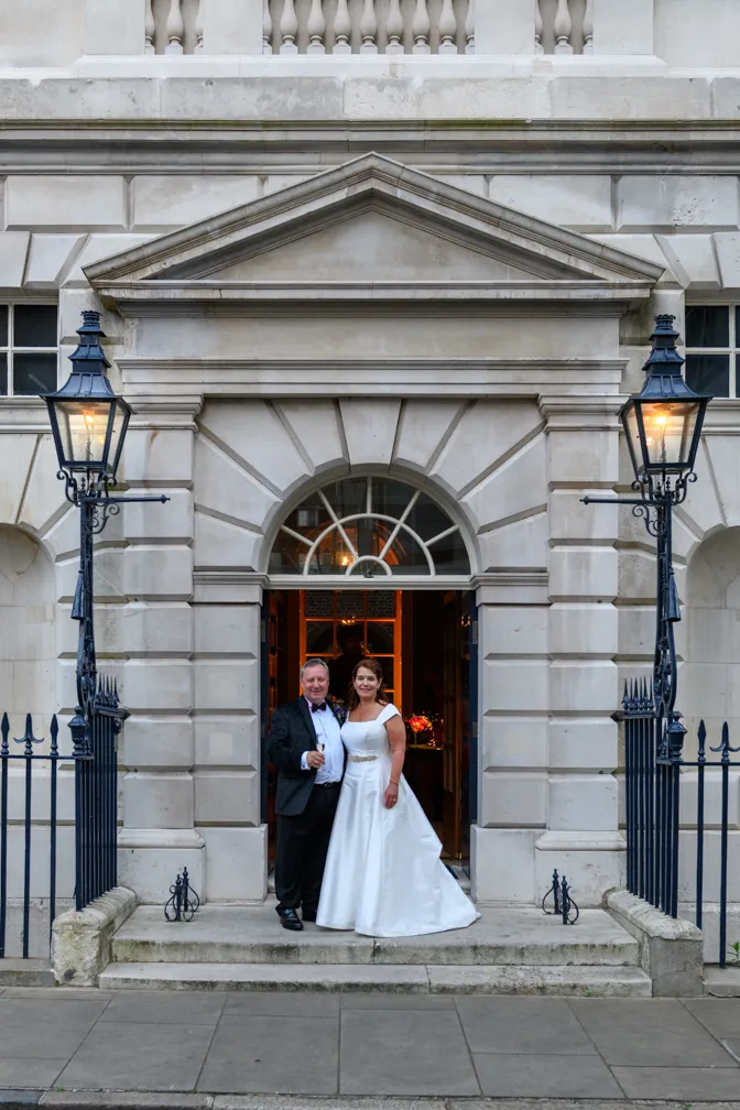 Bride in white gown and groom in black tuxedo standing at the entrance of a stone building with lit lanterns on both sides.