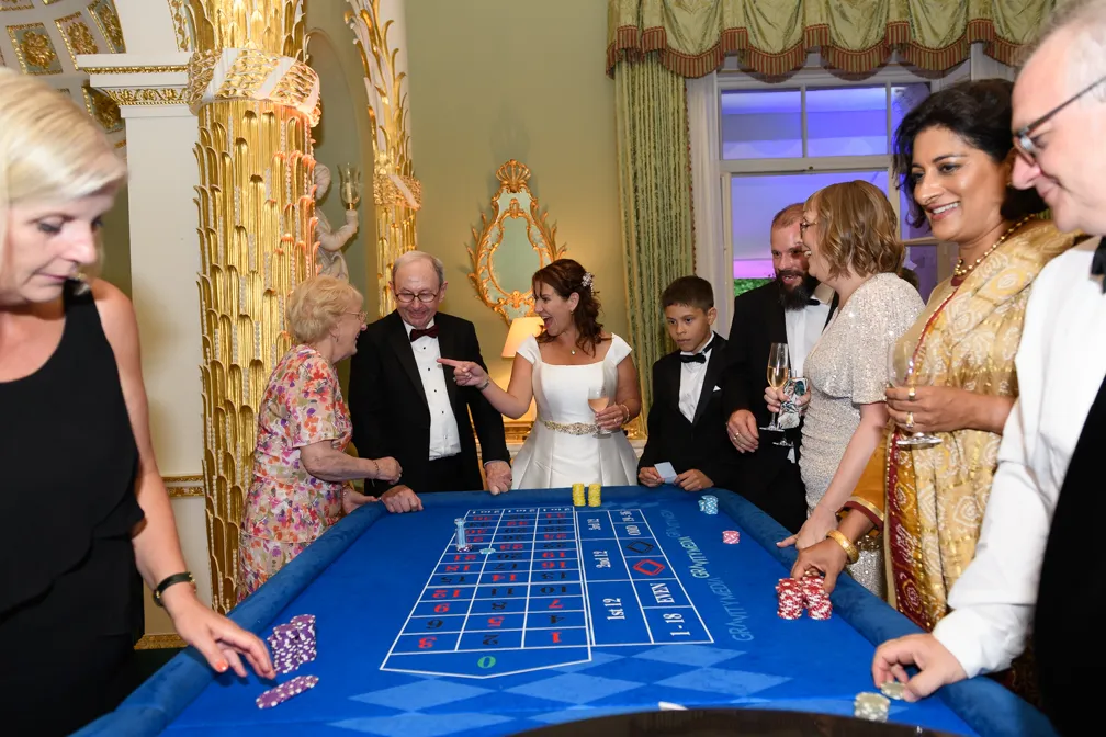 Group of people in formal attire playing at a blue casino roulette table indoors.