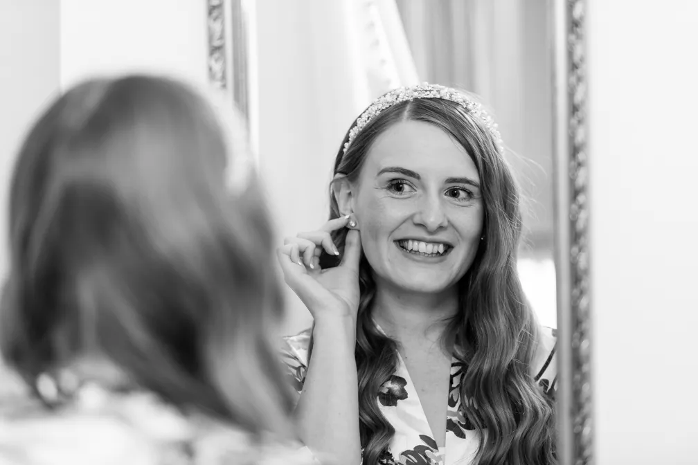 Smiling woman with long hair wearing a decorative headband, adjusting her earring while looking in an ornate mirror.