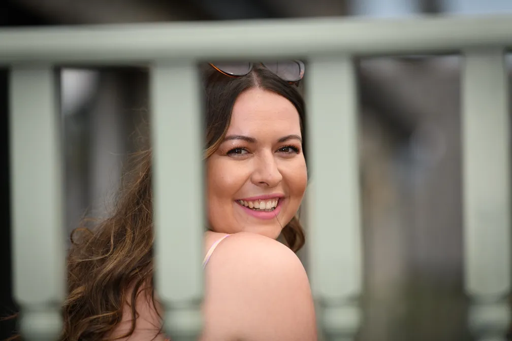 Smiling young woman with long wavy hair looking back over her shoulder through green railings.