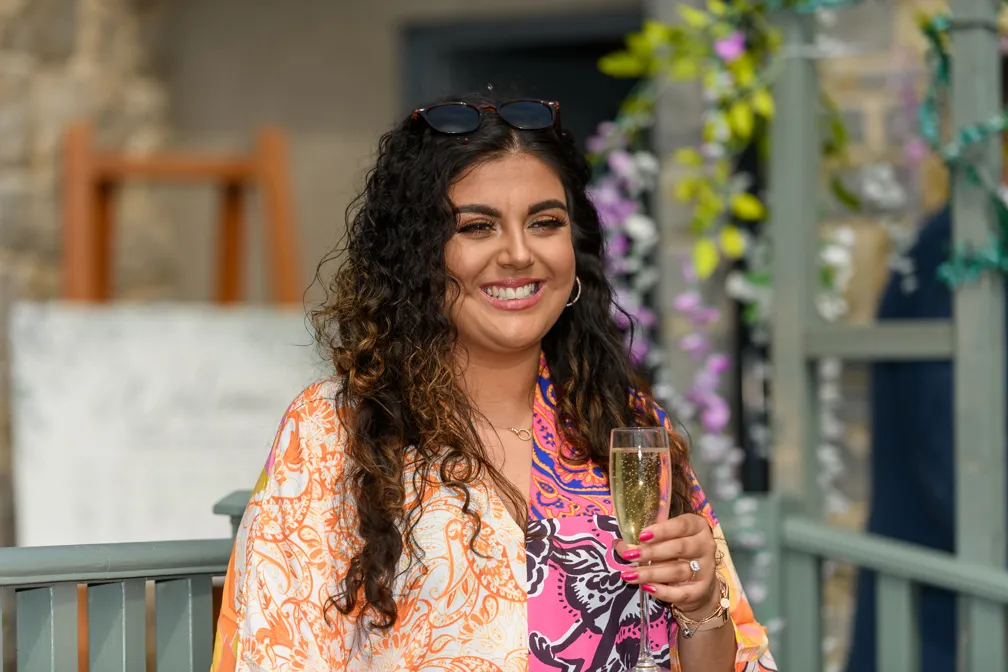 Smiling woman with curly hair holding a glass of champagne, wearing a colorful patterned top and sunglasses on her head.