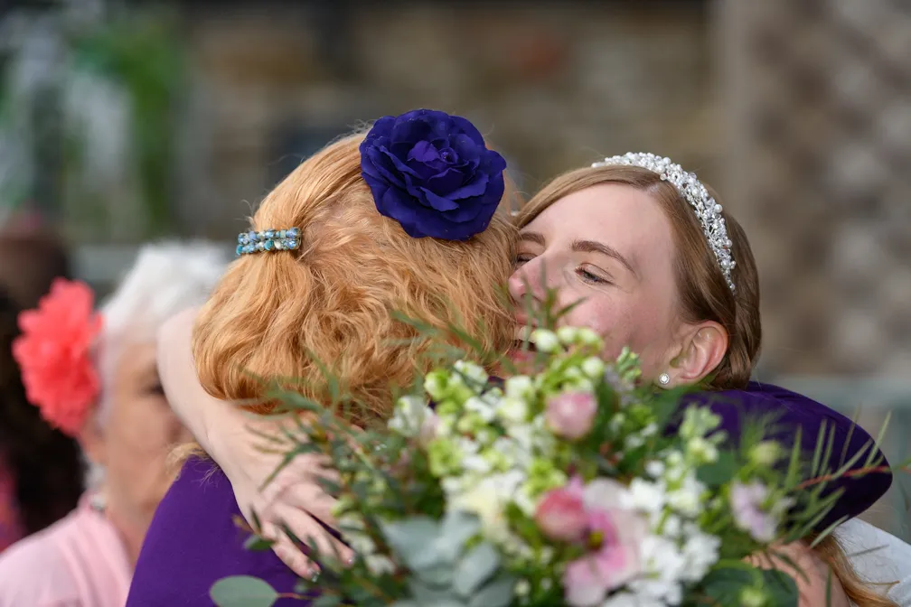 Two women warmly embracing, one wearing a tiara and holding a bouquet with pink and white flowers.