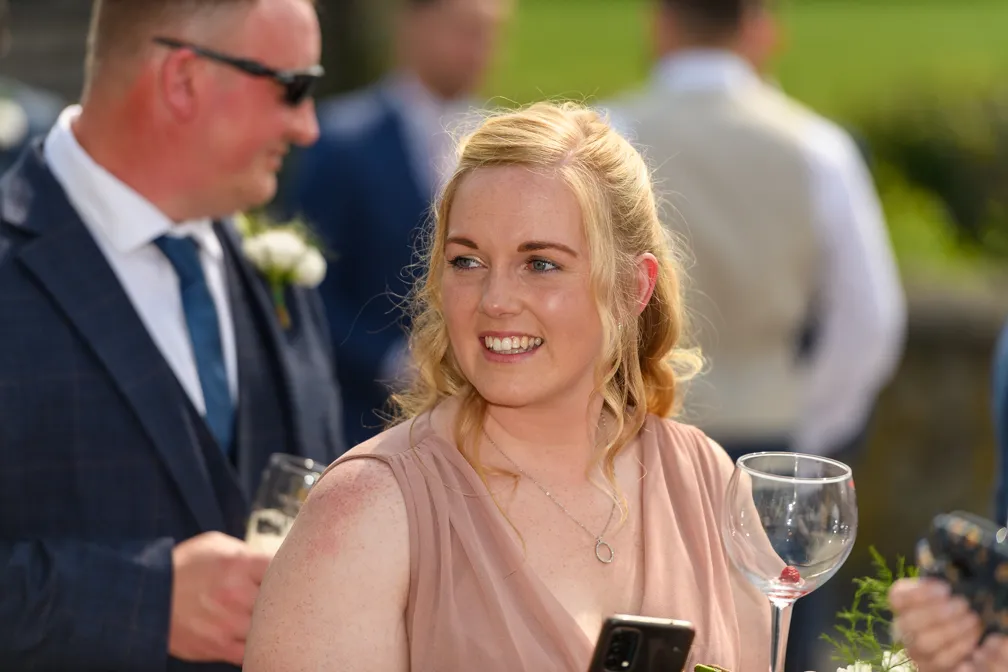 Smiling woman with blonde hair in a peach dress holding a wine glass and a smartphone at an outdoor social event.