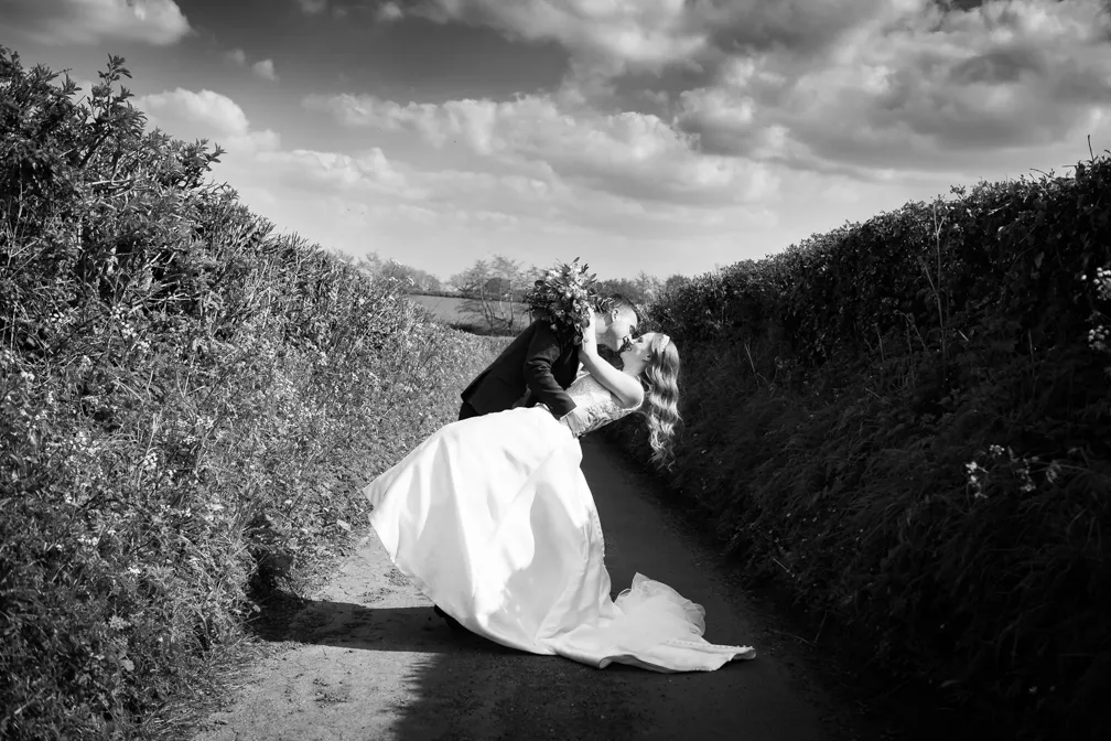Black and white photo of a bride and groom sharing a kiss on a narrow country path surrounded by tall hedge walls.