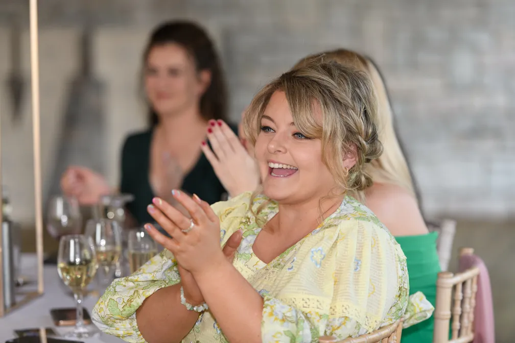Smiling woman with braided hair and a yellow floral dress clapping at a formal event with blurred guests in the background.