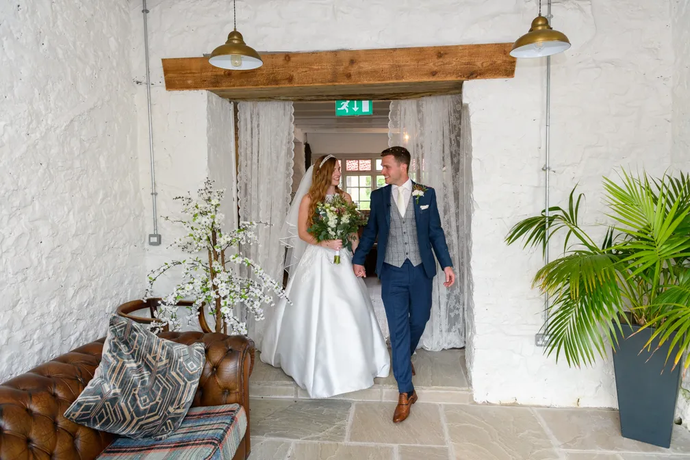 Bride in a white gown holding a bouquet and groom in a blue suit walking hand in hand through a stone doorway with white walls.