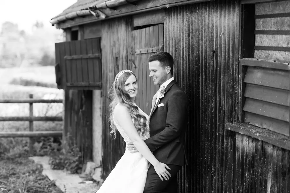 Black and white photo of a bride and groom standing close together outside a rustic building, smiling and embracing.