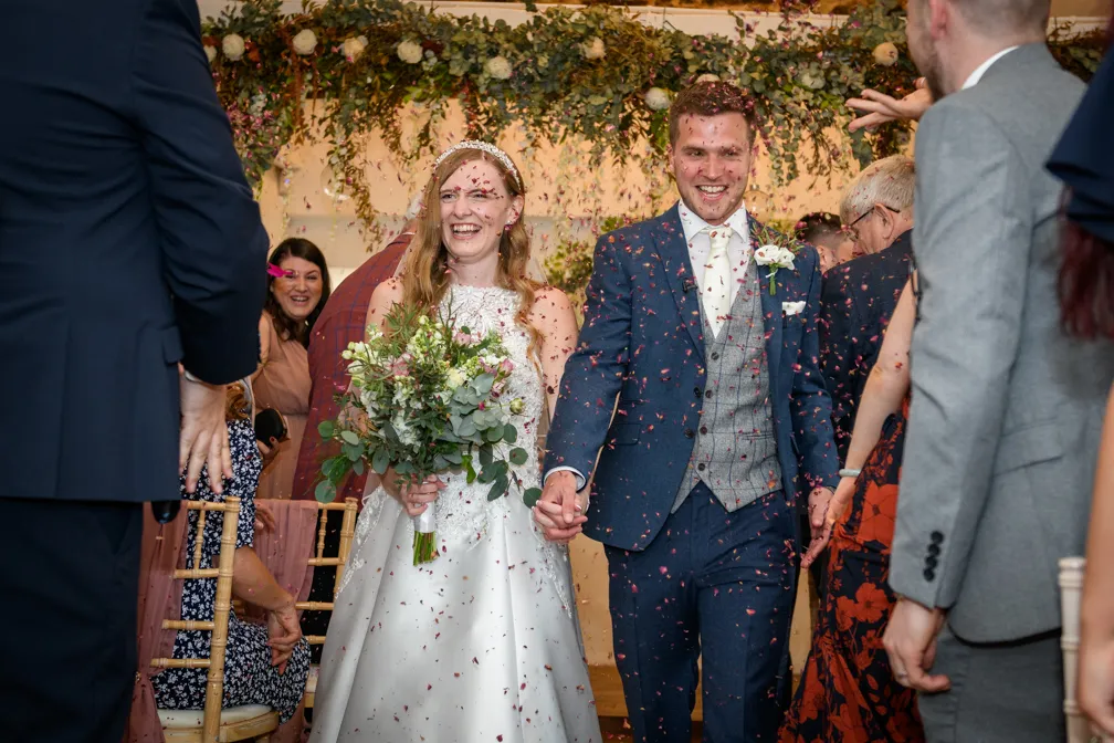 Bride and groom smiling and walking hand in hand down an aisle decorated with greenery as guests throw flower petals.
