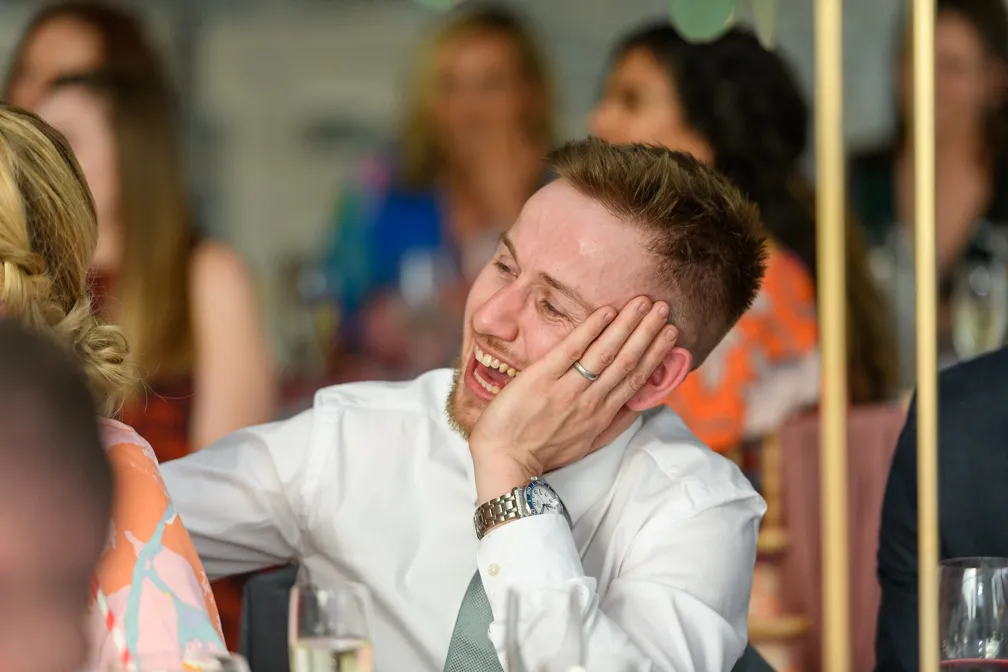 Smiling man in white shirt and tie resting his head on his hand at an event with blurred people in the background.