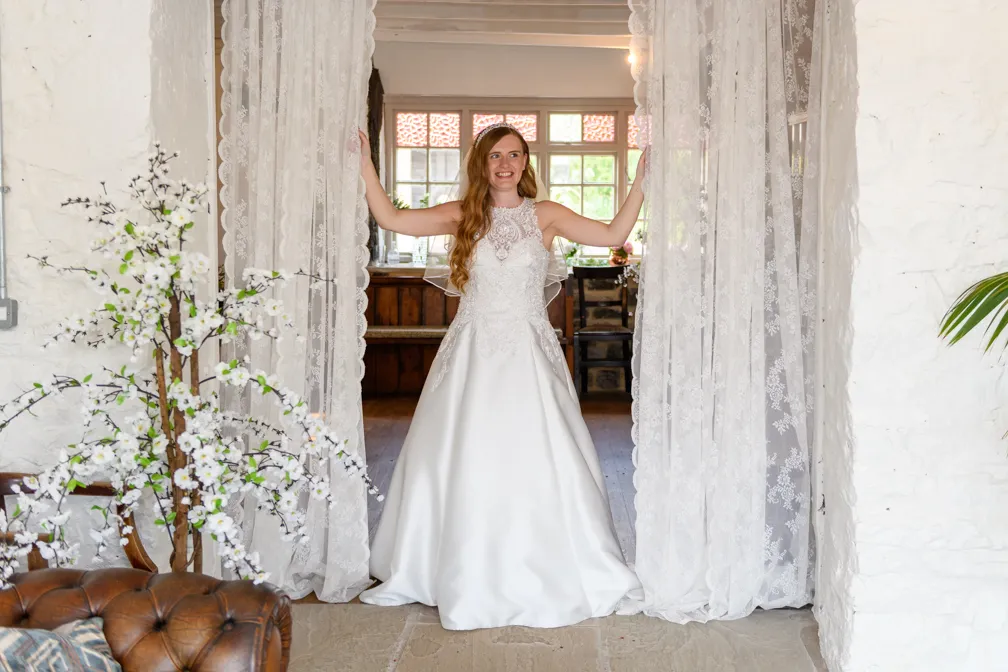 Smiling bride in a white wedding gown standing between lace curtains in a rustic room with wooden furniture and floral decor.