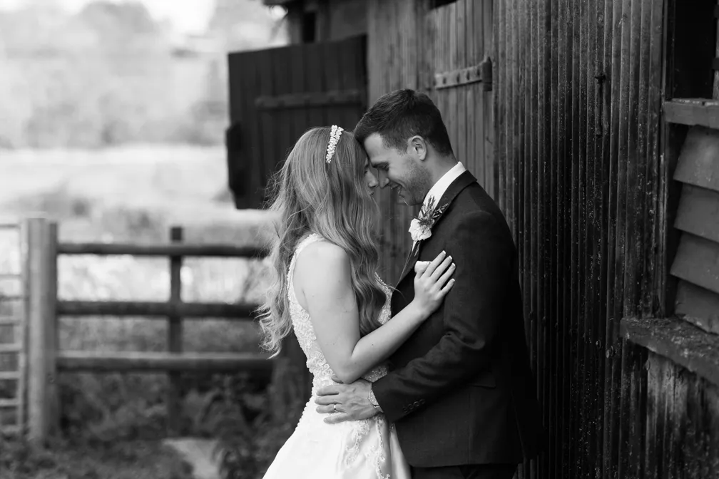 Bride and groom in wedding attire embracing and touching foreheads outdoors near a rustic wooden building.