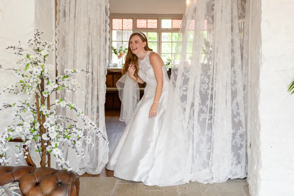 Smiling bride in white lace wedding dress standing behind sheer lace curtains in a bright room with floral decor.