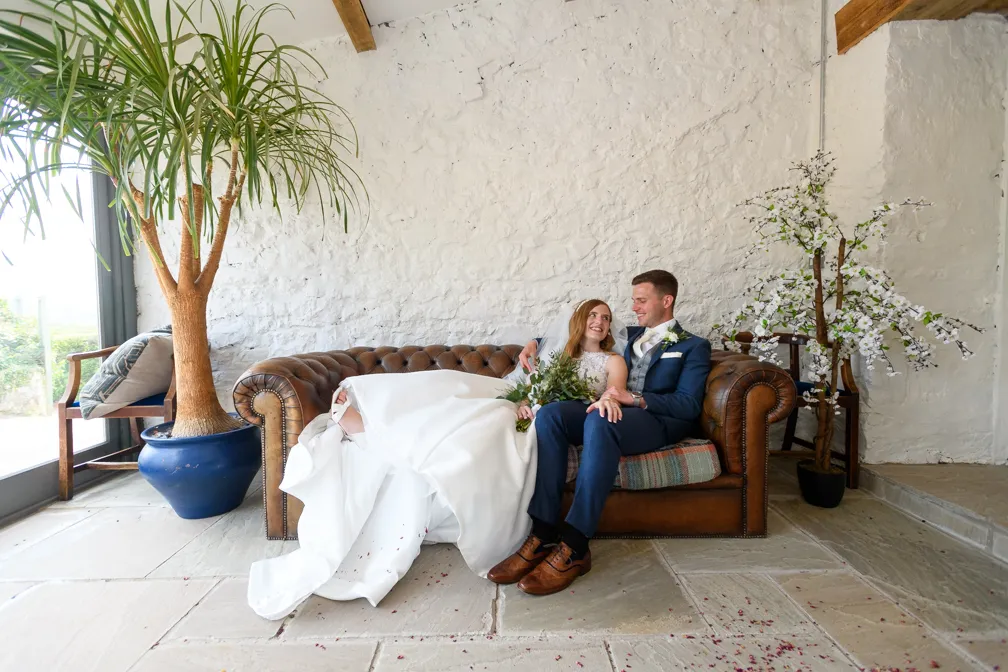 Bride in white wedding dress and groom in blue suit sitting closely on a brown leather couch, smiling at each other indoors with potted plants nearby.