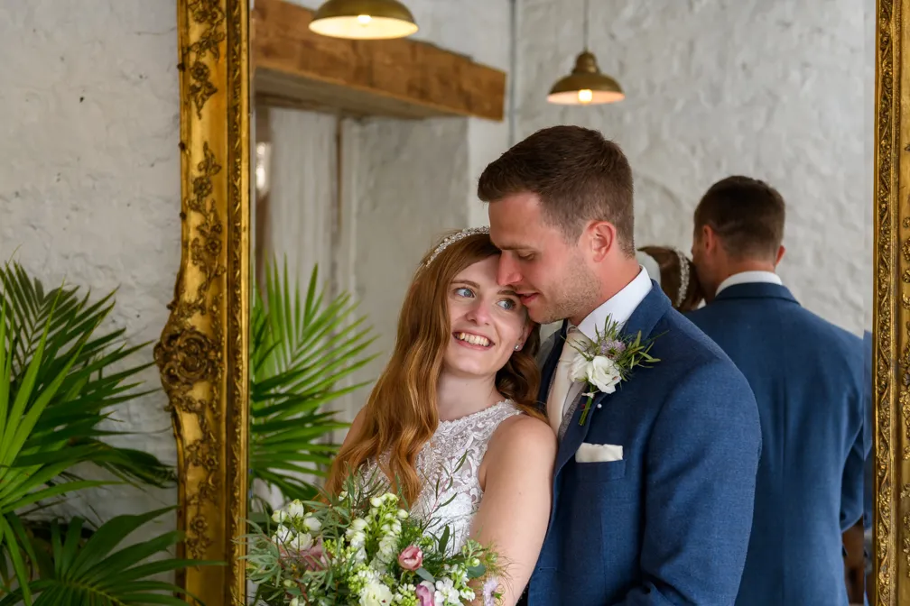 Bride and groom embracing smiling next to a large ornate gold mirror with green plants in the background.