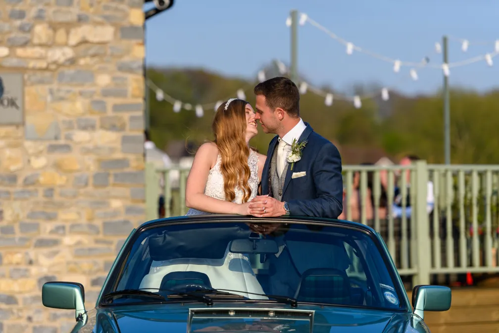 Bride and groom smiling and holding hands while sitting on the back of a black convertible car during their wedding day.