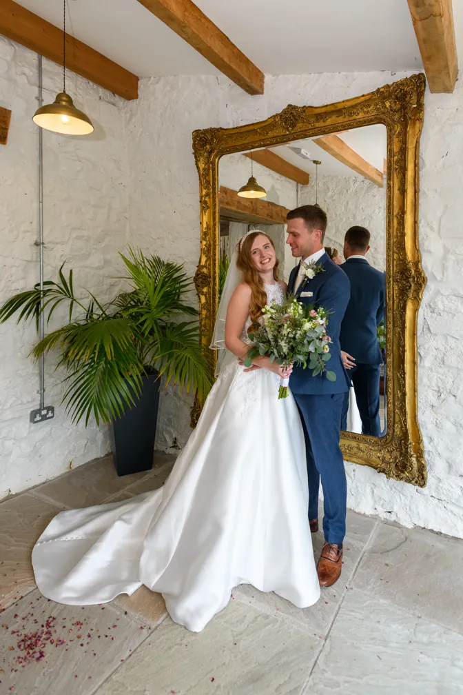 Bride in white gown and groom in blue suit posing together with bouquet in front of a large ornate gold mirror in rustic room.